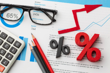 una calculadora, lentes de marco negro y varios lápices sobre documentos con gráficos de barras y porcentajes. Destacan una flecha roja ascendente y símbolos de porcentaje