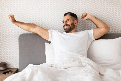 un hombre con barba en la cama, estirando los brazos con una sonrisa radiante tras despertar