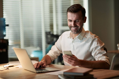 un hombre sonriente trabajando en un escritorio de madera, utilizando simultáneamente una laptop y su smartphone.
