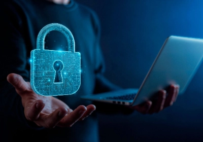 Una figura masculina sostiene una laptop abierta con una mano mientras sobre la otra palma flota un holograma brillante de un candado digital con circuitos integrados.