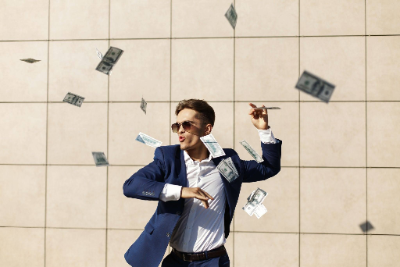 hombre con traje azul y gafas de sol celebrando con entusiasmo mientras caen billetes de dólares a su alrededor.
