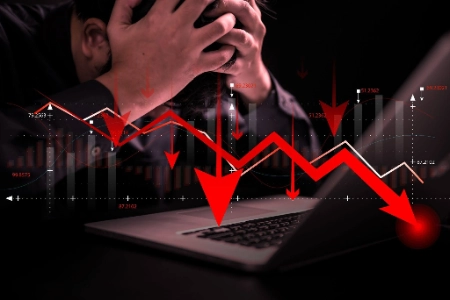 hombre que se cubre el rostro con las manos en un gesto de profunda desesperación frente a una computadora portátil. Sobre la escena se superpone un gráfico financiero con marcadas líneas rojas y flechas que apuntan hacia abajo, simbolizando una caída drástica del mercado o pérdidas económicas severas.
