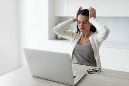 mujer joven sentada frente a su computadora portátil con un gesto de frustración o agotamiento, sosteniendo su cabeza con ambas manos
