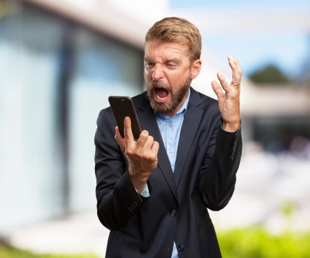 Um homem de terno grita com uma expressão de extrema frustração e raiva enquanto olha para o celular, gesticulando agressivamente com uma das mãos.