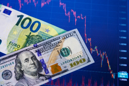 Un billete de 100 dólares y uno de 100 euros colocados en diagonal sobre una pantalla digital azul, la cual muestra un gráfico financiero de líneas rojas en fuerte caída.