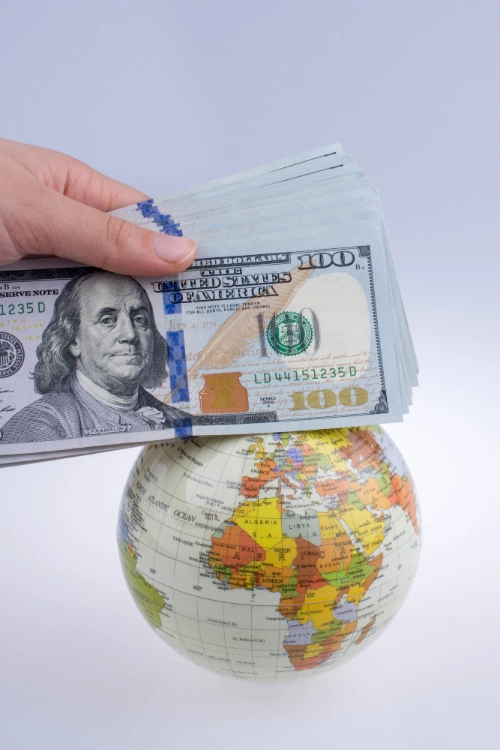 Una foto de plano medio vertical que muestra una mano sosteniendo un fajo de billetes de cien dólares ($100) estadounidenses apoyados encima de un pequeño globo terráqueo