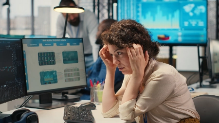 Uma mulher com expressão preocupada e estressada encara seus monitores. As telas exibem gráficos e estatísticas que sugerem um colapso do mercado ou uma crise financeira iminente.