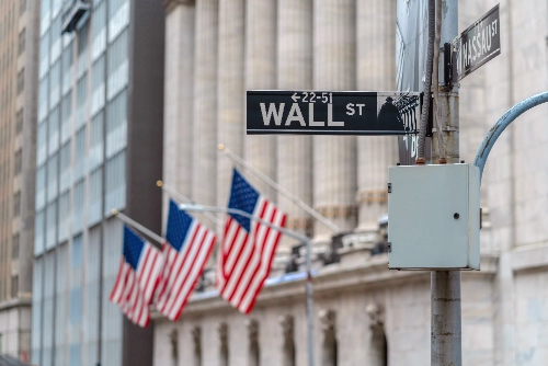 Un primer plano enfocado en el letrero metálico de la calle "Wall St", con varias banderas de los Estados Unidos colgadas