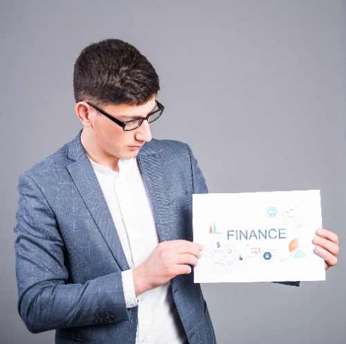Un joven con saco gris y gafas sostiene y mira un cartel blanco que tiene la palabra "FINANCE" impresa junto a varios pequeños iconos gráficos y diagramas sobre un fondo gris liso.