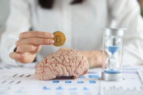 Imagem de grande plano de uma mulher de negócios a segurar uma moeda Bitcoin sobre uma réplica de um cérebro humano, com uma ampulheta azul à direita.