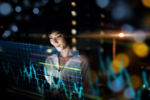 Una mujer analiza gráficos financieros holográficos en azul que flotan frente a ella por la noche. El fondo muestra las luces desenfocadas de una ciudad (bokeh), creando una atmósfera tecnológica y futurista.