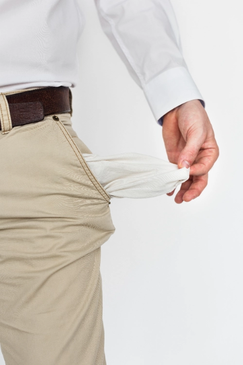 Un hombre vistiendo una camisa blanca y pantalones beige saca hacia afuera el bolsillo delantero de sus pantalones para mostrar que está completamente vacío.