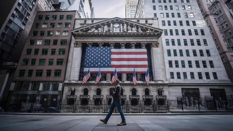 Un transeúnte camina frente al edificio de la Bolsa de Valores de Nueva York (NYSE), cuya fachada neoclásica luce una gran bandera de Estados Unidos entre sus columnas.