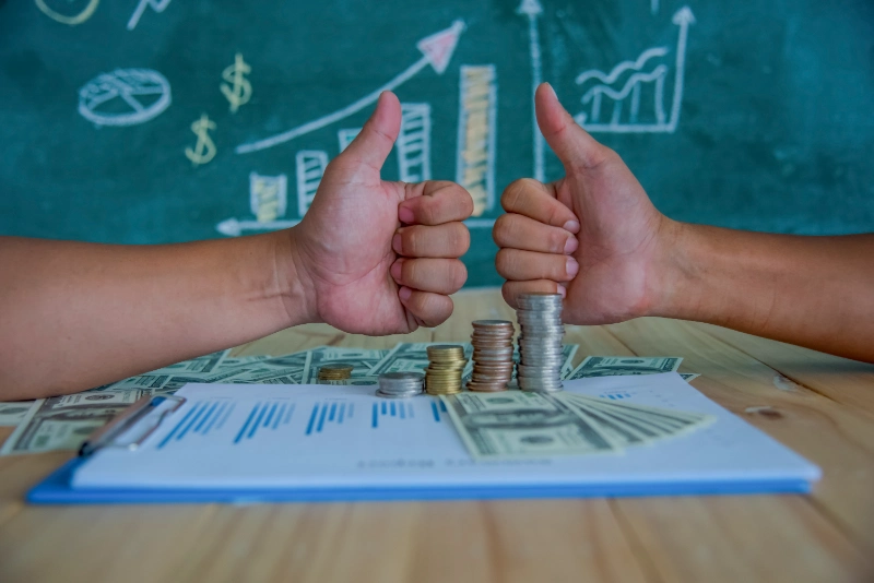 Dos manos haciendo el gesto de pulgar arriba sobre un escritorio con dinero, informes financieros y una pizarra con gráficos de crecimiento de fondo.