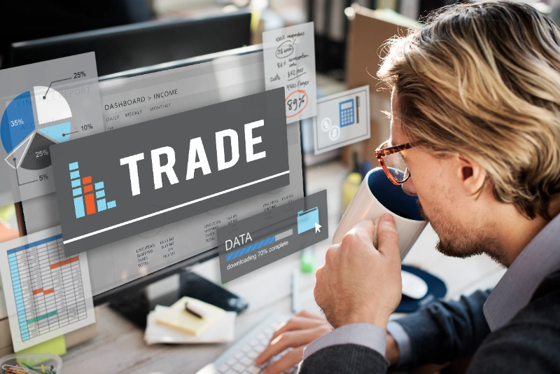 Analista bebiendo café mientras examina datos de comercio y gráficos financieros en la pantalla de su computadora, representando el intercambio comercial y el análisis económico.